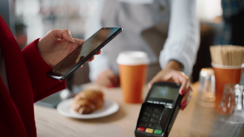 Close Up of a Feminine Hand Holding a Smartphone with an NFC Payment Technology Used for Paying for Take Away Coffee in a Cafe. Customer Uses Mobile to Pay for Latte Through a Credit Card Terminal. Royalty-Free Stock Footage #1062437266