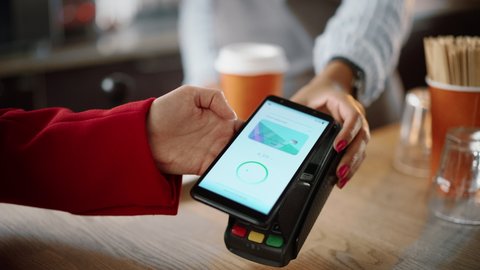 Close Up of a Feminine Hand Holding a Smartphone with an NFC Payment Technology Used for Paying for Take Away Coffee in a Cafe. Customer Uses Mobile to Pay for Latte Through a Credit Card Terminal.