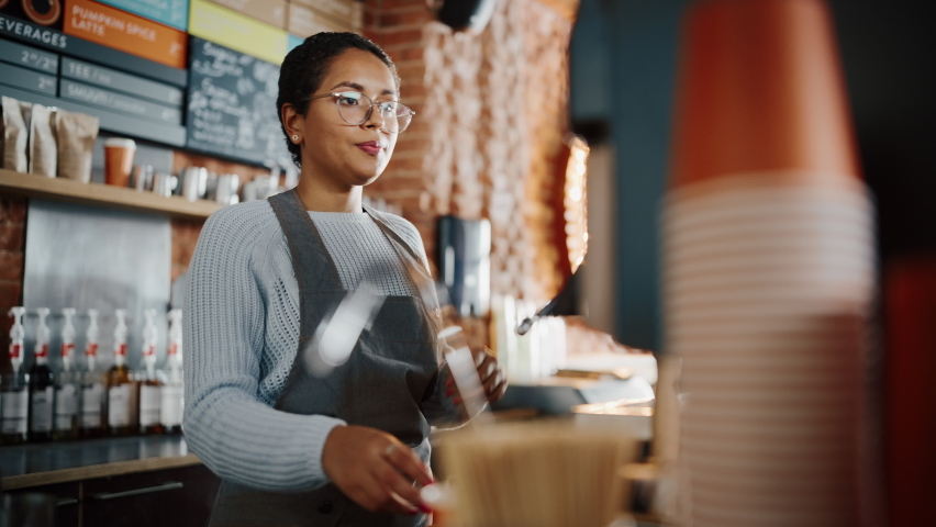 Beautiful Latin American Female Barista with Short Hair and Glasses is Making a Cup of Tasty Cappuccino in Coffee Shop Bar. Male Cashier Works at a Cozy Loft-Style Cafe Counter in the Background. Royalty-Free Stock Footage #1062437314