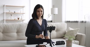 Smiling attractive millennial asian korean mixed female blogger recording make up routine video on smartphone, showing beauty products and tools, recommending cosmetics, modern marketing concept.