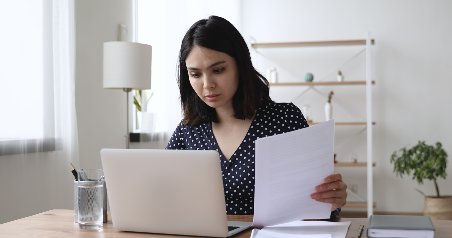 Focused happy millennial asian multiracial businesswoman working on computer with documents, analyzing paper reports, feeling satisfied with research results, distant workday in home office concept. Royalty-Free Stock Footage #1062438403