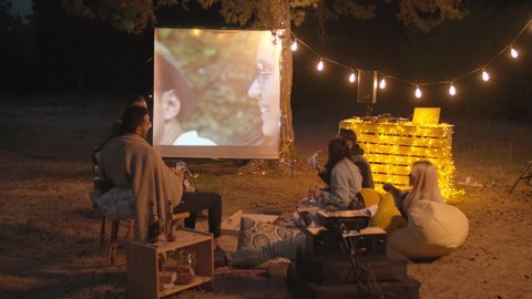 Friends having movie night together watching films on cinema screen outdoors in evening eating pizza and drinking beer