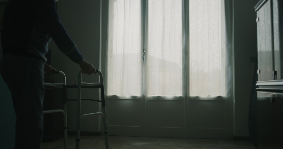 Cinematic shot of an elderly man is walking on stilts towards a window and opening a curtain to look outside . Concept of life, healthcare, retirement, disability, boarding house Royalty-Free Stock Footage #1062441691
