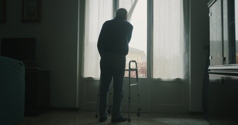 Cinematic shot of an elderly man is walking on stilts towards a window and opening a curtain to look outside . Concept of life, healthcare, retirement, disability, boarding house