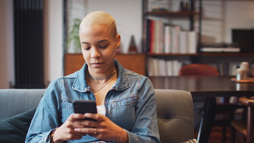 Smiling young woman sitting on sofa at home looking at mobile phone - shot in slow motion