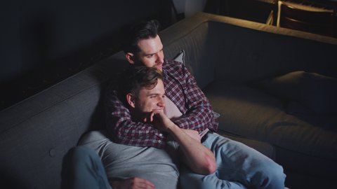 Loving same sex male couple lying on sofa at home hugging and laughing as they watch evening TV together - shot in slow motion
