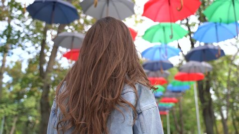 Low angle back view of excited unrecognizable long haired female in denim jacket raising arms and jumping while having fun under hanging colorful umbrellas in autumn park