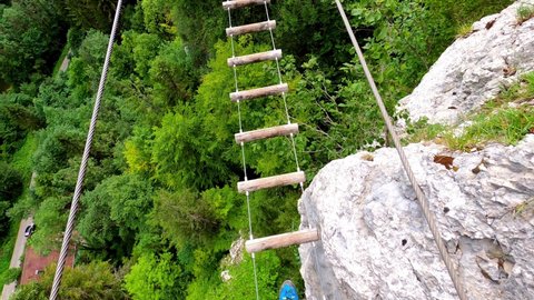 Am man walking on a suspension bridge made out of pieces of wood. Underneath is a forest. Its the via ferrata "ladders of death" in France.