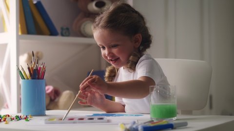 Happy cute little girl preschooler enjoys painting process, dipping paintbrush into watercolor, smiling joyfully. Kid having fun during art lesson.
