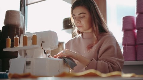 A young woman sews clothes, sitting in a light workshop at a table with a sewing machine. She smiles and enjoys creative work. Medium shot. Fashion designer, fashion business.