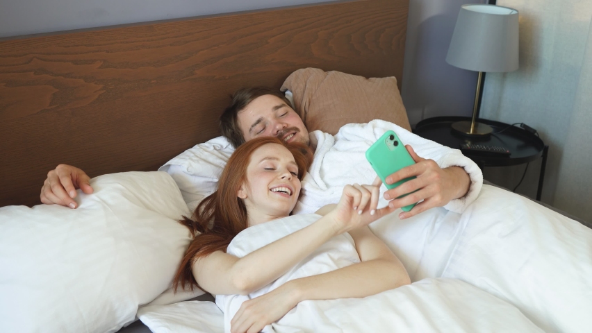 Young happy couple makes online breakfast caucasians lying in hotel room bed