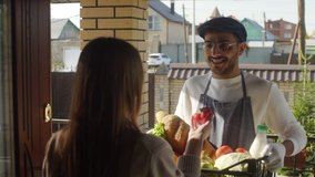 Young woman opening door of her house, smelling bell pepper and taking crate with fresh produce, bread and dairy product from cheerful male farmer while receiving farm-to-door delivery - Powered by Shutterstock - Get 15% off with code: PIKWIZARD15