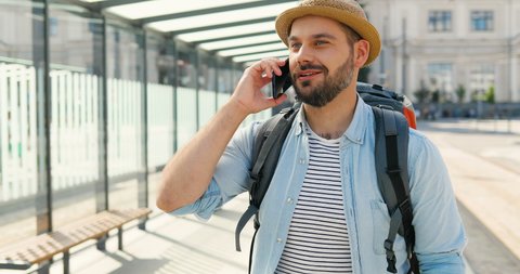 Caucasian young handsome man tourist in hat with backpack talking on telephone and smiling at train station on summer day. Attractive male traveller speaking on mobile phone at bus stop Cellphone call