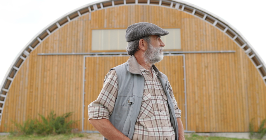 Portrait shot of Caucasian senior handsome man in hat standing outdoors at big wooden shed, looking at side and turning to camera. Old male farmer at his land. Dolly shot.