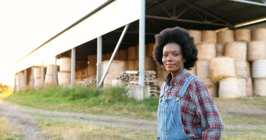 Portrait of beautiful African American young woman farmer standing in field at shed with hay stocks and smiling to camera. Pretty female with smile at farm. Countryside concept.