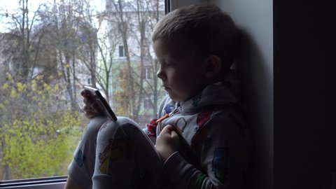 four year old boy with smartphone on windowsill. View of autumn Park and street space for text. Danger of internet