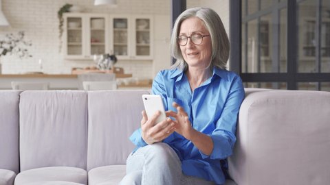 Excited happy mature old 60s woman customer winner holding smartphone using mobile app winning online lottery bid, celebrating success, receiving gift voucher on cell phone sitting on couch at home.
