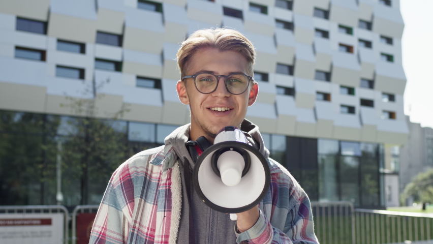 Young Guy Speaks Into Loudspeaker On The Street. Cute Blond Guy With Headphones Around His Neck Raises His Hand Up And Has Fun Talking. He Smiles. Behind Him The Building.