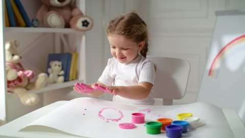Cute happy little girl preschooler uses hands covered in pink paint to create palm prints on big sheet of paper. Kid spending time at home with pleasure.