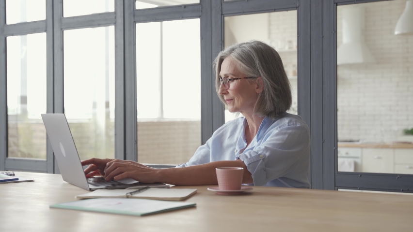 Senior older professional mature business woman using laptop computer sits at workplace desk. Happy mid aged employee 60s grey-haired businesswoman executive working typing on pc at home from office. | Shutterstock HD Video #1062468226