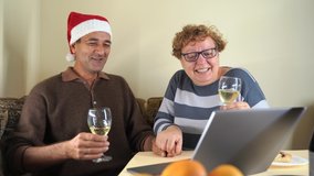 Happy mature husband and wife with glasses of champagne sitting at table with laptop and proposing toast for friends during online meeting and Christmas celebration at home