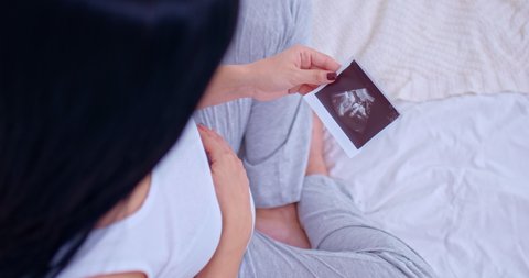 Happy motherhood, a pregnant woman looks at a photo of an ultrasound scan of her baby and strokes her belly. Close-up, a pregnant woman communicates with an embryo, strokes her belly with her hand