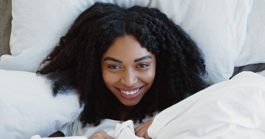 Happy playful african american woman peeking out from her blanket in bed and smiling to camera, top view, close up | Shutterstock HD Video #1062472045