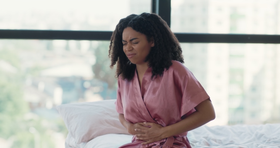Sick black woman suffering from stomach ache sitting in bed, holding belly, feeling abdominal or menstrual pain in morning. Gastritis, diarrhea and painful periods concept