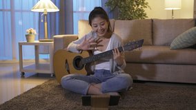 cheerful taiwanese young lady holding guitar is live streaming at home. asian female cover singer greeting her fans waving hi in front of pad and starting to play songs on instrument.
