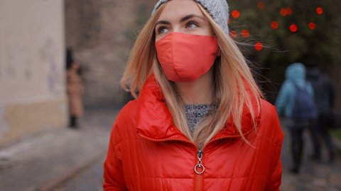 Beautiful blonde in knitted hat and warm red jacket spending free time on fresh air. Pretty woman wearing red medical mask while walking at city center.