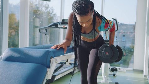 Strong and healthy. Young african american woman athlete is doing hard physical exercise in gym, doing dumbbell traction, treadmill and exercise bike in the background. Healthy lifestyle, sports