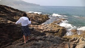 A young girl with a camera walks along the rock going into the sea. Waves crash on stones