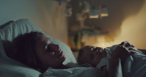 Cinematic shot of an young peaceful  mother is sleeping with her newborn baby boy  on bed in nursery at night. Concept: sweet dreams,childhood,life,motherhood maternity, authenticity