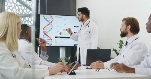 Front view of good-looking confident experienced bearded medical worker holding a presentation on interactive screen for cvalified diverse male and female colleagues