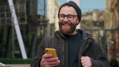 Close up view of bearded male person in 30s looking at phone screen and smiling while walking at street. Handsome guy in glasses chatting in social media while using smartphone