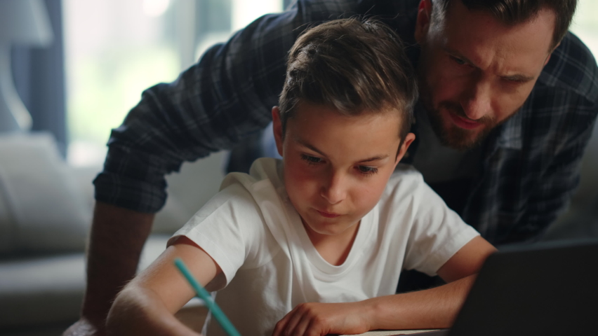 Closeup father helping son doing homework in living room. Smiling child and parent looking computer screen at home. Positive man standing behind back of boy during distance learning online indoors. | Shutterstock HD Video #1062495406