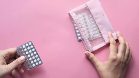 women's hand putting birth control pills in a small bag on pink background 
