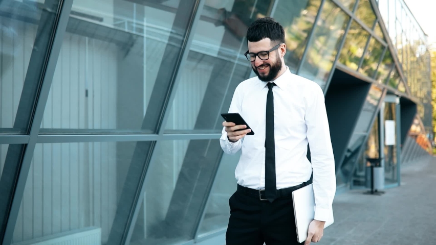  young modern businessman using mobile phone while walking outdoors near glass building of business center Royalty-Free Stock Footage #1062500866
