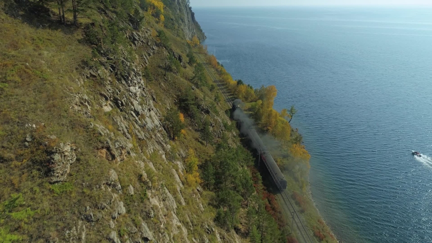 Old steam locomotive train rides along shore blue Lake Baikal Russia Circum-Baikal Trans-Siberian Railway. Epic autumn rocky natural landscape. Sunny. Open space horizon. Aerial forward flight film