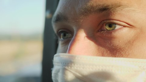 Close up of green-eyed middle-aged man in face mask looks out the window and at camera on a bus ride. Travel safety during coronavirus pandemic quarantine concept