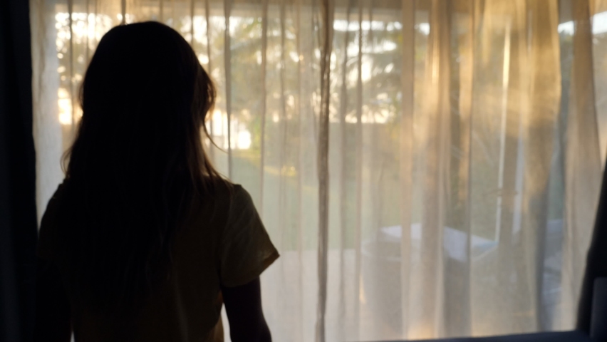 Slow motion: Young woman walking towards room window opens curtains and walks into the garden. Beach bungalow, woman on vacation  Royalty-Free Stock Footage #1062504319