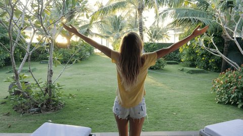 Slow motion: Young woman walking towards room window opens curtains and walks into the garden. Beach bungalow, woman on vacation 