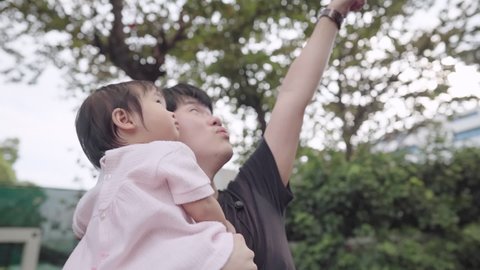 young Asian dad and his adorable cute infant baby girl looking up to the sky inside the park standing under trees, child care parenting bonding, children innocence curiosity, father and daughter
