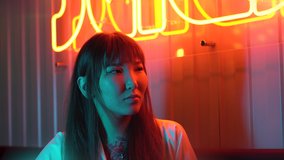 Portrait of a young beautiful Asian woman against the background of neon lights. Fast food restaurant with Asian cuisine. Large red sign with neon hieroglyphs.