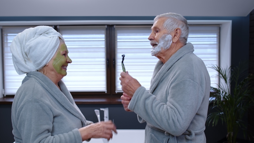 Old senior couple man and woman in bathrobe are dancing in bathroom having fun laughing enjoying leisure time and music. Elderly grandmother with facial green mask and grandfather with shaving foam