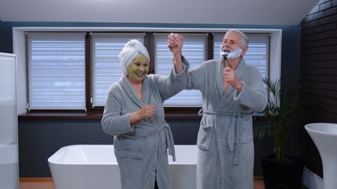 Old senior couple man and woman in bathrobe are dancing in bathroom having fun laughing enjoying leisure time and music. Elderly grandmother with facial green mask and grandfather with shaving foam