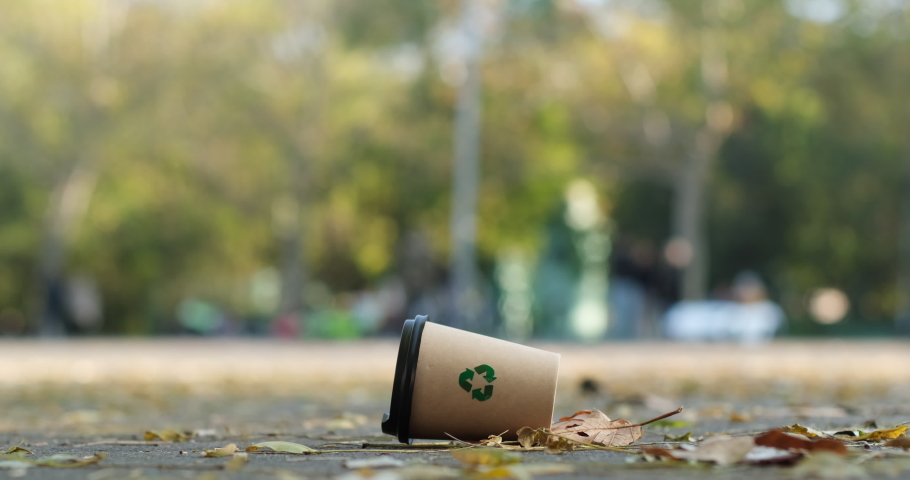 A paper cup with the "recycle" sign lies among the autumn leaves on the park road against the background of people walking in the park. Recyclable waste lies under people's feet. Royalty-Free Stock Footage #1062513667