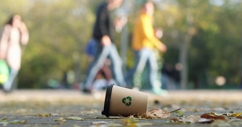 A paper cup with the "recycle" sign lies among the autumn leaves on the park road against the background of people walking in the park. Recyclable waste lies under people's feet.