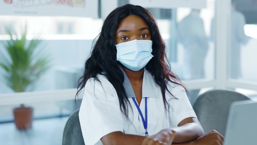 Young afro-american attractive female intern nurse wearing respiratory mask sitting in medical office therapist workspace waiting for patients. Young specialist. Ethnicity. Hospital staff. | Shutterstock HD Video #1062517189
