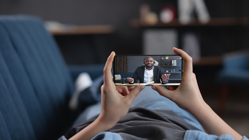 The woman is lying on a Coach in Living Room, watching Webinar, Seminar, holding Video-Call. Young African American Mentor Couch is holding Online Webinar, wearing Suit and Headsets. Working Indoor.